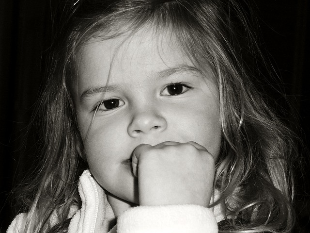 Close-up photo of a young girl looking intently at the camera. Her left hand obscures her mouth. Her long hair is slightly untidy.